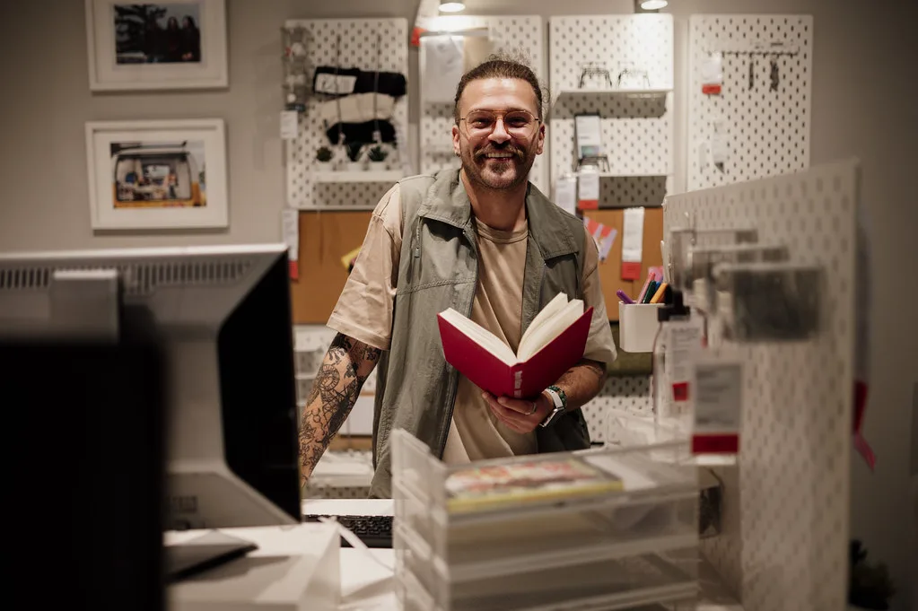 技术服务与办公场景：Man holding a book and smiling in a creative workspace with various supplies in the background